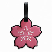 A pink cherry blossom-shaped luggage tag with a black strap, featuring a white stylized flower center and black border on a white background.