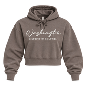 A cropped pullover hoodie in a washed taupe-brown color, featuring 'Washington' and 'DISTRICT OF COLUMBIA' printed in white script across the chest. The hoodie is shown against a plain white background.