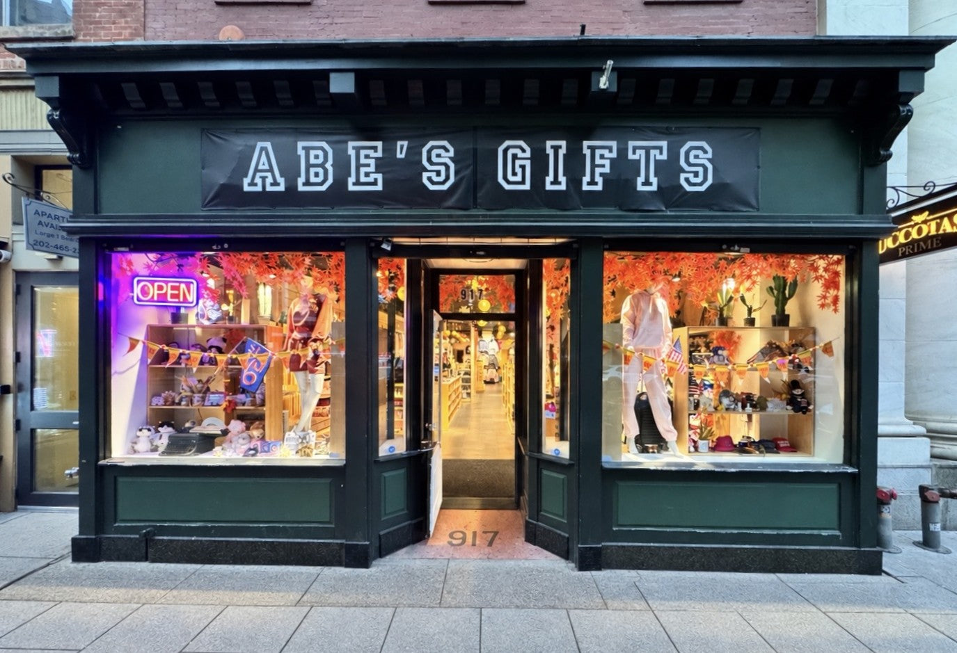 A storefront facade for Abe's Gifts, with a dark green frame, large display windows, an open sign in the window, and mannequins inside. The sign above the entrance reads 'ABE'S GIFTS' in white letters.
