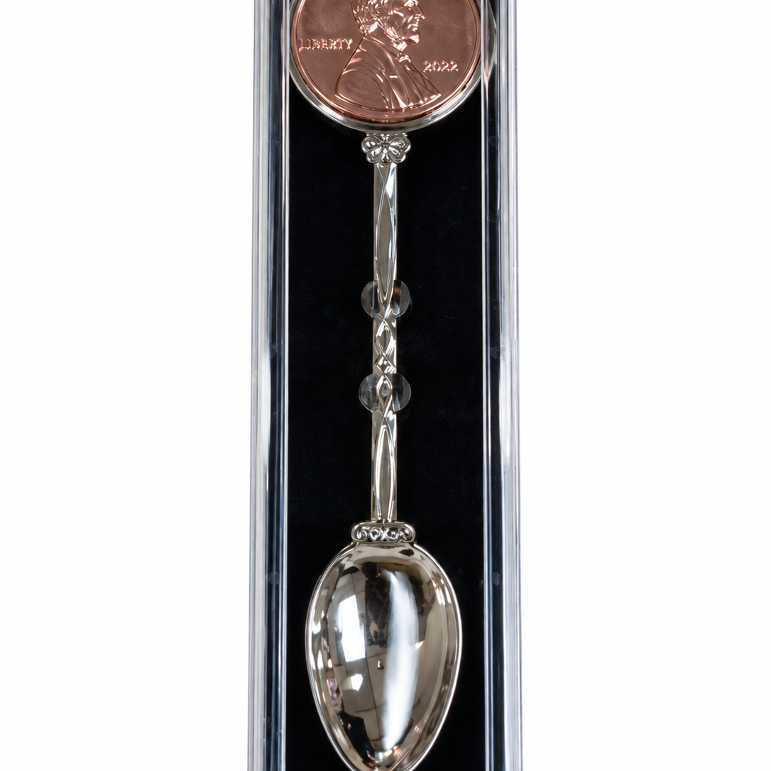 A silver spoon-shaped bookmark with a penny charm, presented inside a clear plastic protective case with a black insert, on a white background.