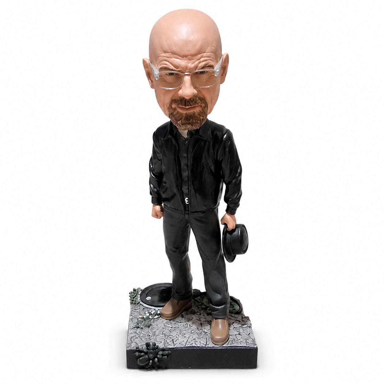 A collectible bobblehead figurine of Walter White from Breaking Bad, standing on a textured gray base with a black hat in one hand, dressed in a black jacket and pants with brown shoes.