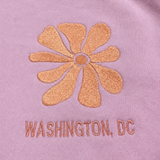 Close-up of a lavender-purple fabric surface with a metallic rose-gold embroidered flower and the text 'WASHINGTON, DC' stitched below it.
