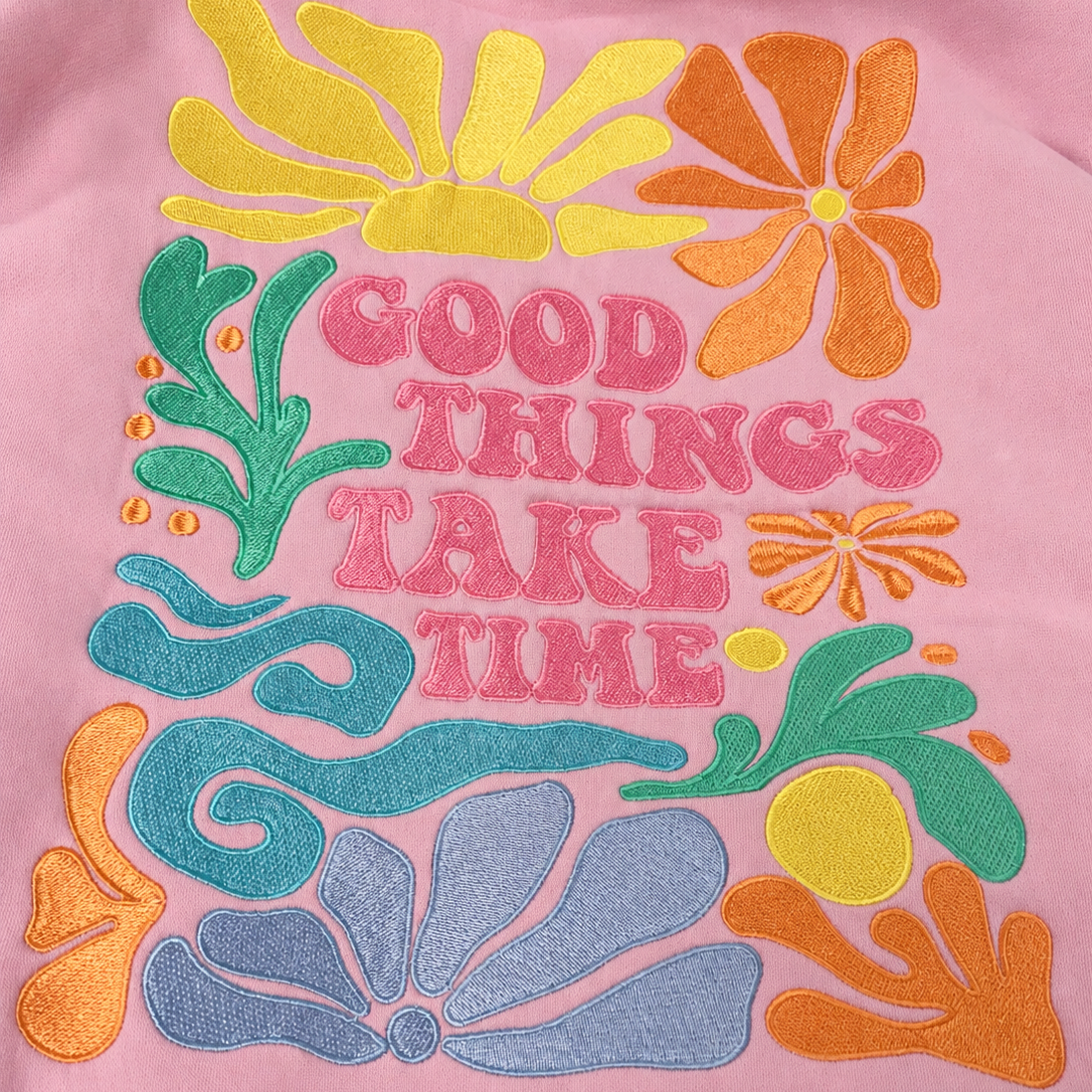 A close-up of a pink sweatshirt featuring a colorful retro embroidered graphic with the text 'GOOD THINGS TAKE TIME' in pink, surrounded by yellow, orange, green, and blue floral motifs.