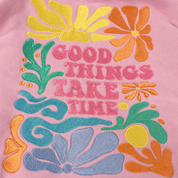 A close-up of a pink sweatshirt featuring a colorful retro embroidered graphic with the text 'GOOD THINGS TAKE TIME' in pink, surrounded by yellow, orange, green, and blue floral motifs.