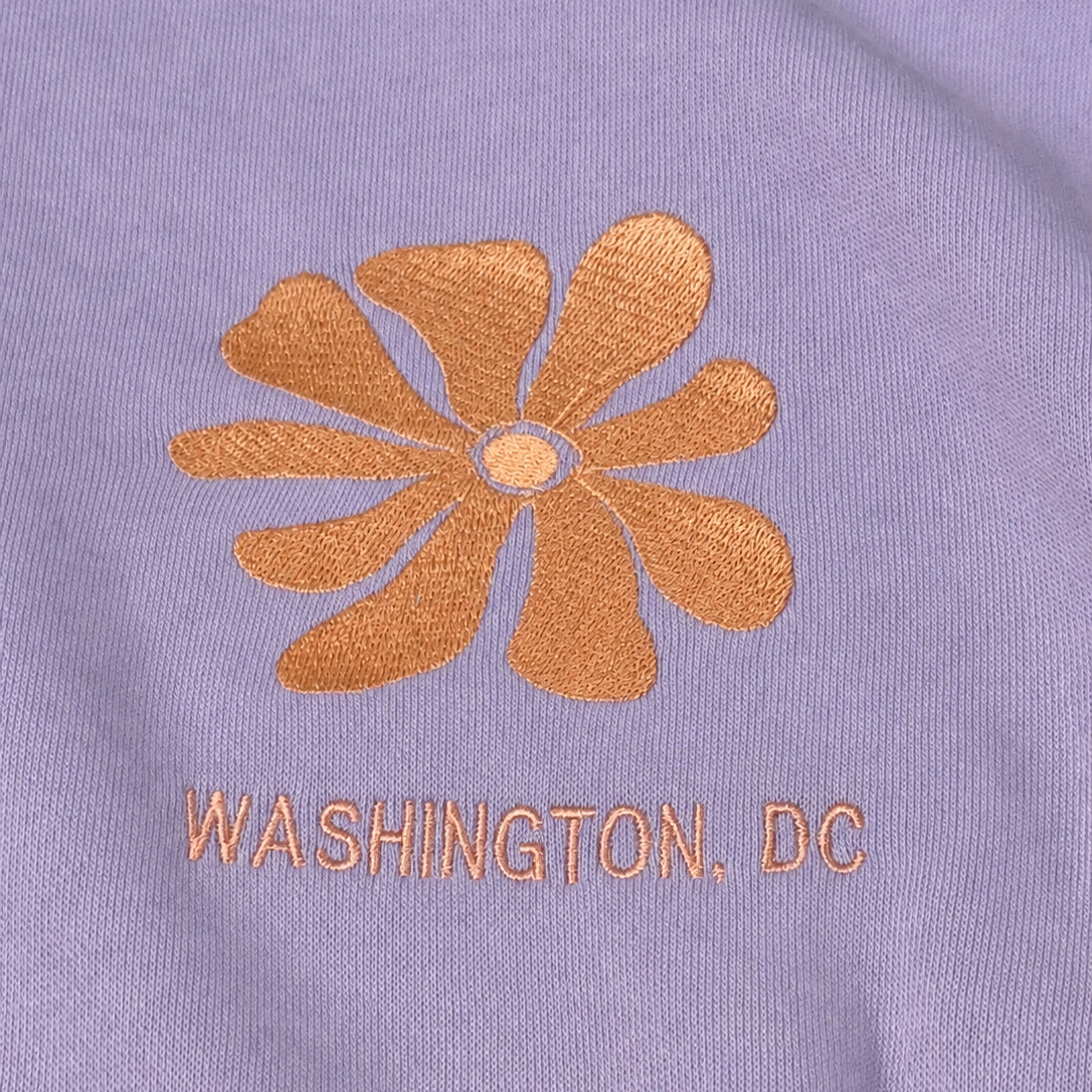 Close-up of a lavender-purple fabric surface with a metallic copper embroidered flower and the text 'WASHINGTON, DC' below it.