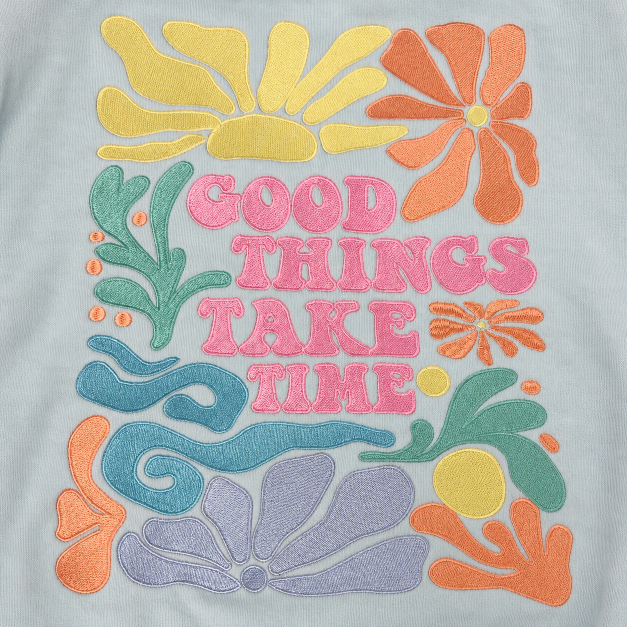 A close-up of a light gray fabric featuring colorful embroidered retro-style floral motifs and the phrase 'GOOD THINGS TAKE TIME' in pink lettering.