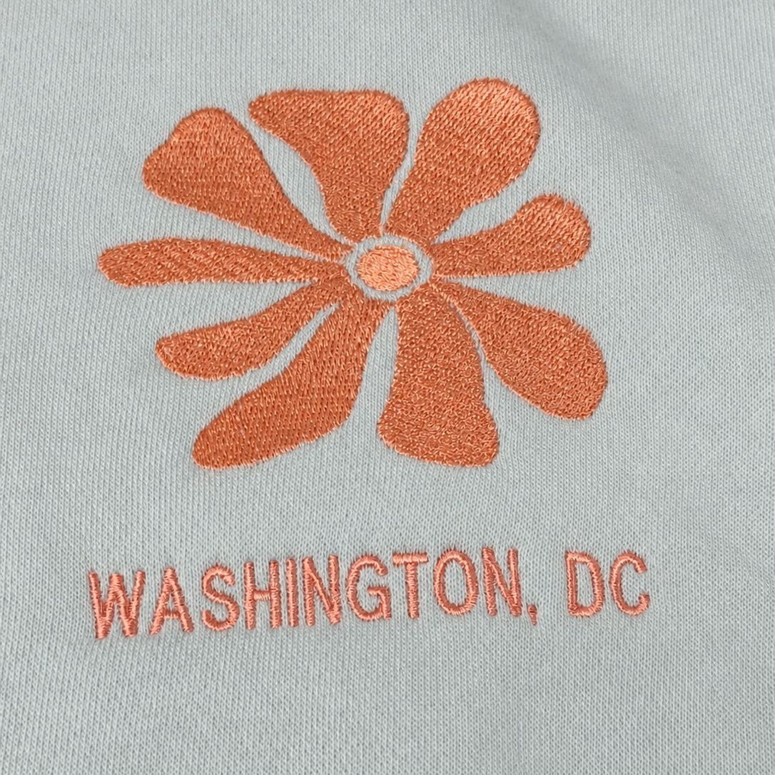 Close-up of a light gray fabric surface with an orange embroidered flower and the text 'WASHINGTON, DC' in matching orange embroidery.