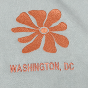 Close-up of a light gray fabric surface with an orange embroidered flower and the text 'WASHINGTON, DC' in matching orange embroidery.