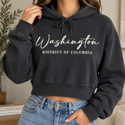 A woman wearing a dark charcoal gray cropped hoodie with 'Washington District of Columbia' printed in white script and block lettering on the front, paired with light blue jeans.