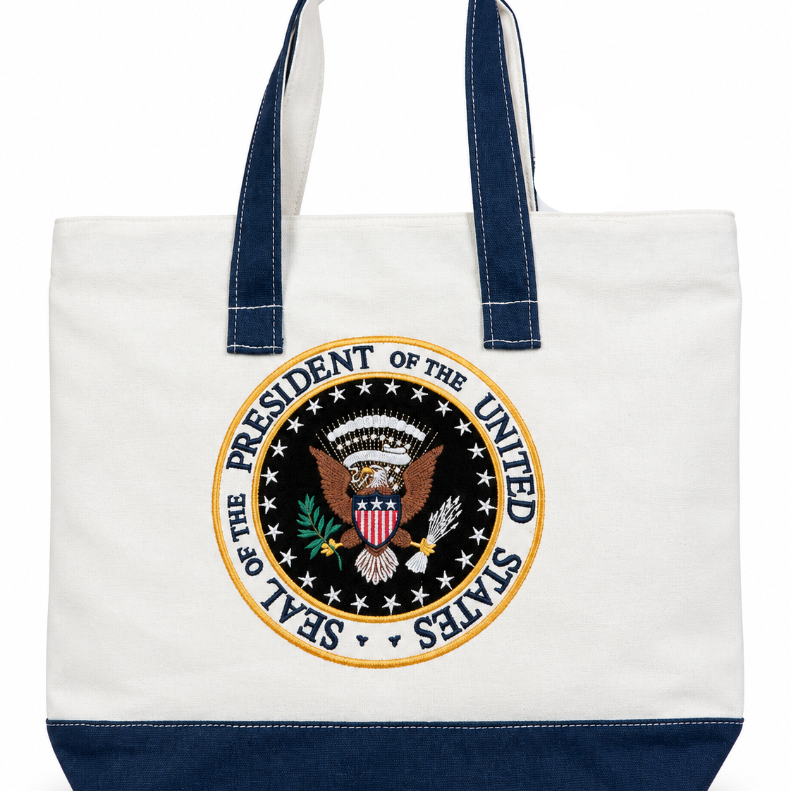 A canvas tote bag with a white body, navy blue handles and bottom trim, featuring a large circular Seal of the President of the United States printed on the front.